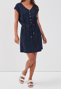 Abito navy blue a maniche corte con scollatura a V, chiusura a bottoni, vita regolabile con lacci e texture liscia, abbinato a sandali bianchi.