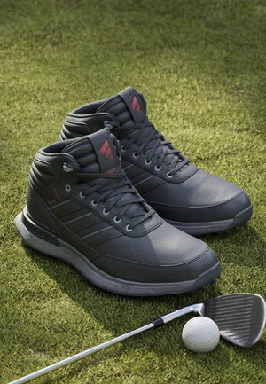 Schwarze Leder-Golfstiefel mit strukturiertem Obermaterial, Design mit drei Streifen und roten Akzenten, kombiniert mit einem Golfschläger und einem Ball auf dem Gras.