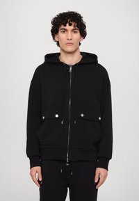 Sudadera negra de tela gruesa con cremallera frontal, dos bolsillos en el pecho con broches y cordones en la capucha. Textura suave, corte relajado.