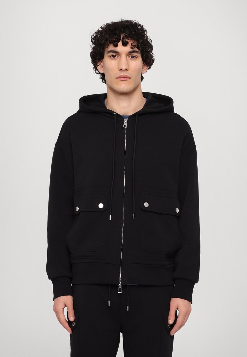 Sudadera negra de tela gruesa con cremallera frontal, dos bolsillos en el pecho con broches y cordones en la capucha. Textura suave, corte relajado.