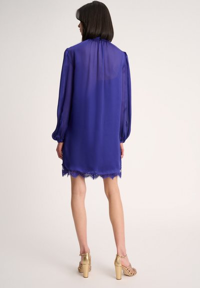 Paarse chiffon jurk met lange, volumineuze mouwen en kantafwerking aan de zoom. Draag je met gouden hoge hakken. Achteraanzicht.