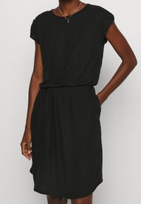 Schwarzes, kurzärmeliges Kleid aus weichem Material, mit einem lockeren Schnitt, gerafftem Tailleneinsatz und seitlichen Taschen. Einfacher Rundhalsausschnitt und glatte Textur.