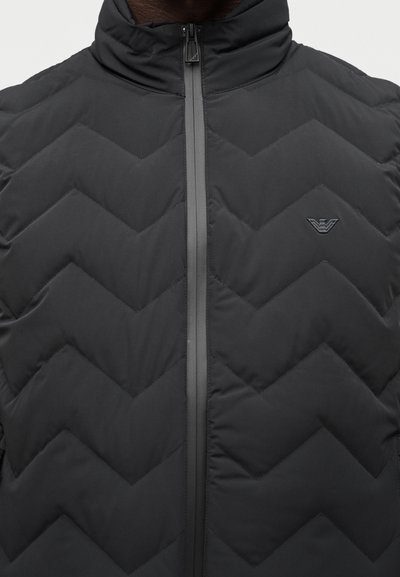 Μαύρο καπιτονέ μπουφάν με ραφές σε σχήμα βέλους, ψηλό γιακά, κλείσιμο με φερμουάρ και μικρό κεντητό λογότυπο στο στήθος.