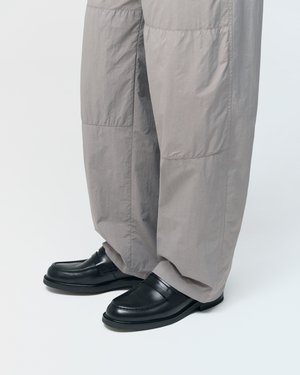 Mocassins en cuir noir portés avec un pantalon ample gris clair, debout sur une surface blanche.