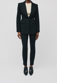 Musta räätälöity bleiseri terävillä läpyillä, yksittäisellä napilla, ja kahdella etutaskulla, yhdistettynä kapeahelmaisiin mustiin housuihin ja teräviin mustiin kenkiin.