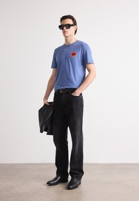 Blue cotton t-shirt with round red logo, paired with black denim pants and black leather boots; holding a black leather jacket.