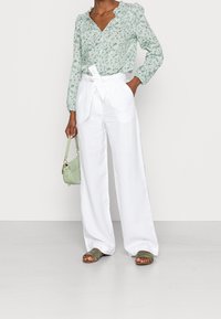 Kvinna som bär en ljusgrön blommig blus, högmidjade vita vida byxor med bälte, gröna sandaler och håller en liten grön handväska.