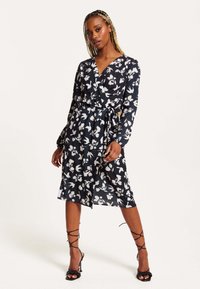 Black wrap dress with white floral pattern, long sleeves, tie belt at the waist, and a knee-length hem. Worn with black high-heeled sandals.
