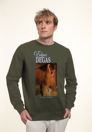 Giovane uomo con felpa oliva che presenta il dipinto di Edgar Degas "Alle scuderie" con un cavallo e un piccolo cane in una scena di stalla.