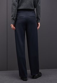 Persona con pantalones de pierna ancha negros, con bolsillos traseros y cinturilla elástica, combinados con un suéter gris oscuro y zapatos negros, de pie en el interior.