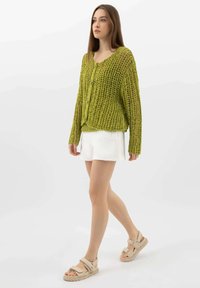 Cardigan verde a maglia con vestibilità ampia, dotato di scollatura a barca e chiusura con bottoni. Abbinato a una gonna bianca e sandali beige.