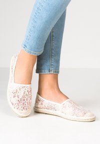 Espadrilles en dentelle de couleur crème, avec un design à mailles ouvertes et une semelle en jute tissé. Associées à un jean léger à revers bleu clair.