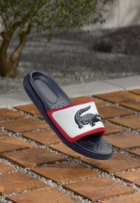 Marineblå slide sandal med en hvid og rød rem, der har et sort logo. Tekstureret sål, afslappet design med en fleksibel gummibyggelse.