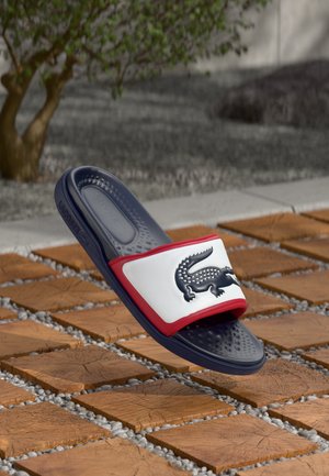 Sandale à glissière bleu marine avec une sangle blanche et rouge, comportant un logo noir. Semelle intérieure texturée, design décontracté avec une construction en caoutchouc flexible.