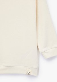 Kremowy sweter dresowy o prążkowanej fakturze, z długimi rękawami i małą metką z wizerunkiem misia na dolnej krawędzi. Miękki, casualowy design.