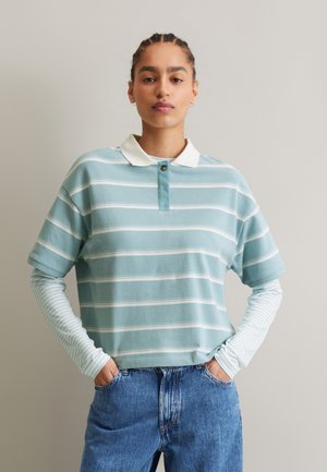 Hellblau gestreiftes Poloshirt mit einem weißen Kragen und einem Knopfdetails, über einem langärmligen gestreiften Shirt getragen, kombiniert mit blauen Jeans.