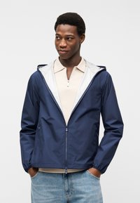 Άνδρας που φοράει μπλε-ναυτικό φούτερ με φερμουάρ και κουκούλα πάνω από μπεζ πόλο μπλουζάκι και ανοιχτόχρωμα τζιν, στέκεται με τα χέρια στις τσέπες μπροστά σε λευκό φόντο.