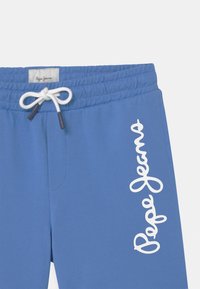 Modré tepláky vyrobené z měkké látky, s elastickým pasem a bílou stahovací šňůrkou a výrazně potištěným bílým logem.