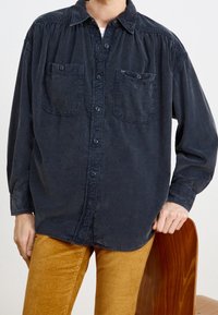 Persona che indossa una camicia button-up blu scuro con due tasche sul petto e pantaloni di corduroy giallo senape, con la mano poggiata sullo schienale di una sedia di legno.