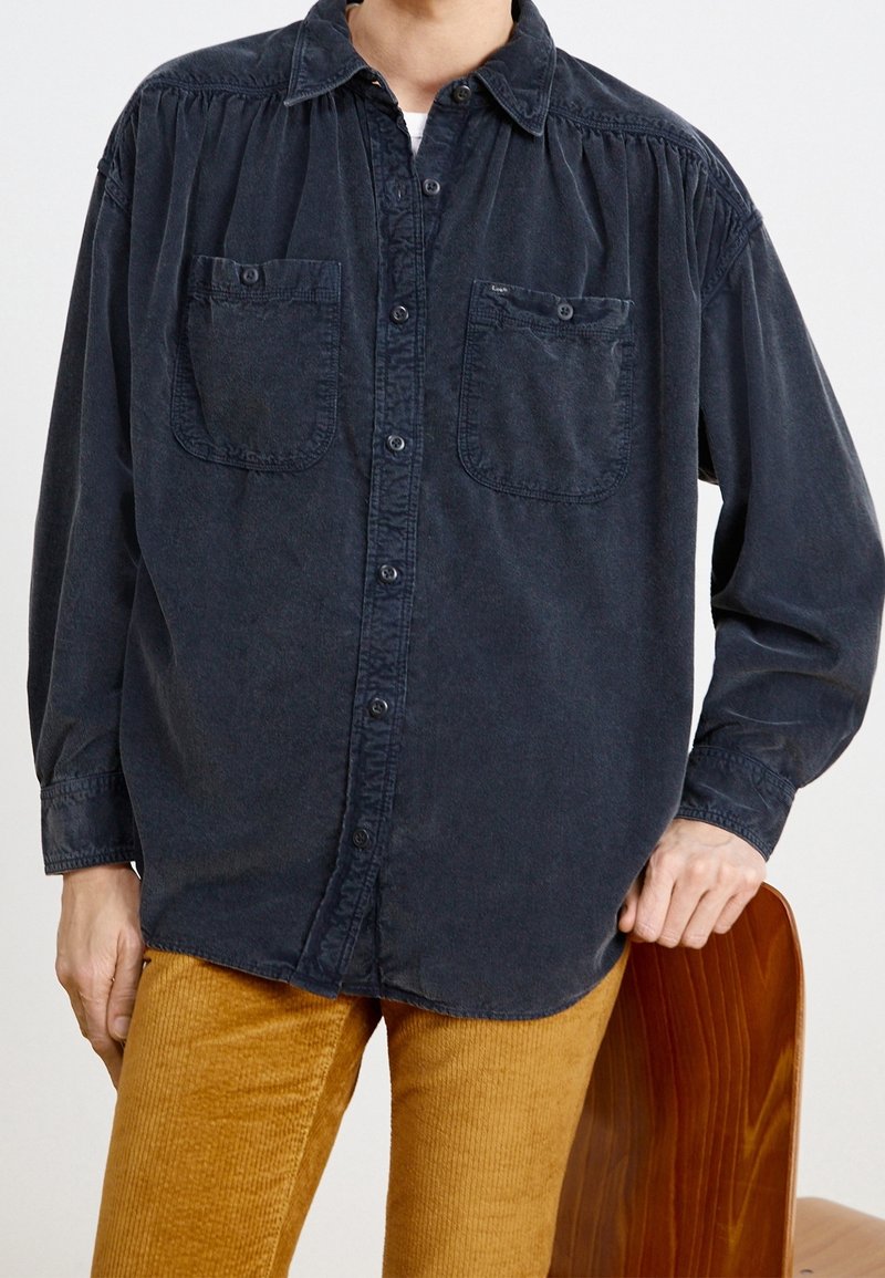 Persona che indossa una camicia button-up blu scuro con due tasche sul petto e pantaloni di corduroy giallo senape, con la mano poggiata sullo schienale di una sedia di legno.
