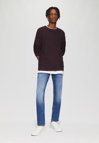 Dunkellila Pullover mit strukturiertem Finish, über einem weißen Hemd getragen; kombiniert mit hellblauen Jeans und weißen Sneakers.