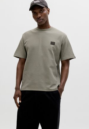 Homme portant un t-shirt beige à manches courtes en tricot gaufré avec une petite écusson sur la poitrine, un pantalon noir et une casquette noire avec un logo.