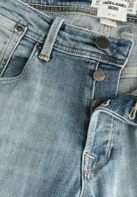 Jeans de denim azul claro con una cintura alta, botones de metal con texto en relieve y costuras naranjas en contraste.