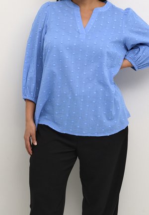 Frau trägt eine hellblaue, strukturierte Bluse mit Dreiviertelärmeln und schwarze Hose, steht mit einer Hand in der Hüfte vor einem einfarbigen Hintergrund.