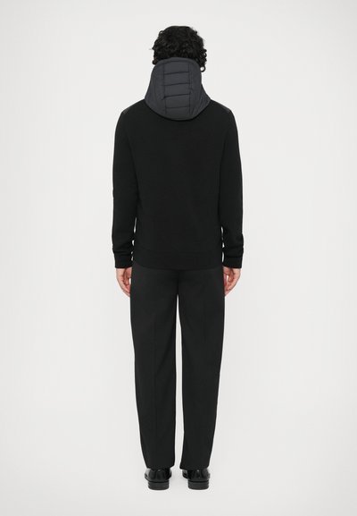 Pulôver preto com capuz acolchoado, apresentando uma textura suave e punhos canelados, combinado com calças e sapatos pretos, visto por trás.