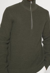 Olivegrüner Strickpullover mit hohem Kragen und silbernem Reißverschluss. Strukturiertes Gewebe mit Rippdetails an den Bündchen und dem Saum.