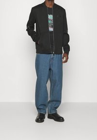 Giacca bomber nera con zip frontale, dotata di due tasche anteriori e di una vestibilità moderna, abbinata a jeans blu larghi e stivali neri.