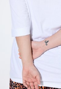 T-shirt blanc à manches retroussées, associé à un bas à motifs orange et noir. Un tatouage est visible sur le poignet de la main derrière le bras.