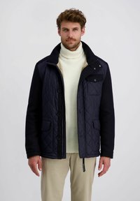 Mann trägt eine marineblaue gesteppte Jacke über einem cremigen Rollkragenpullover und beigen Hosen, steht vor einem schlichten hellen Hintergrund.