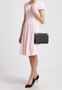 Femme dans une robe rose pâle mi-longue et des talons hauts noirs, tenant un sac à bandoulière noir avec une chaîne dorée.