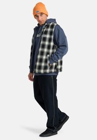 Black and white plaid vest over a blue hoodie, paired with black pants and black shoes. Orange knit beanie adds color contrast.
