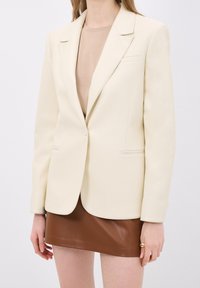 Femme portant un blazer écru sur un haut beige associé à une mini-jupe en cuir marron, debout devant un fond uni.