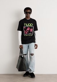 Svart grafisk t-skjorte med grønn tekst, lagdelt over en hvit langermet skjorte; slitte lyseblå jeans; svart totebag; svarte sko.