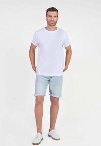 Man staat met handen in zijn zakken, draagt een wit T-shirt, lichte denim shorts en witte sneakers tegen een effen lichte achtergrond.