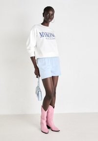 Hvit hettegenser med blå tekst, lyseblå shorts, rosa cowboystøvler og en blå mønstrete handbag. Jevn tekstur, uformelt design.