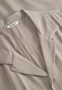 Blusa beige chiaro con una texture morbida, dotata di colletto piegato, pieghe sottili e l'etichetta "GABBA" cucita all'interno del collo.