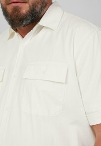 Korte mouw, witte blouse met kraag van een gestructureerde stof, voorzien van twee voorzakken en subtiele verticale strepen.
