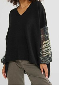 Femme portant un haut noir à col en V avec des manches bouffantes transparentes à motifs et un pantalon vert olive sur un fond uni.