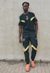 Conjunto de ropa deportiva negra que incluye una camiseta con detalles en verde y amarillo, y pantalones a juego. Zapatillas naranjas brillantes complementan el conjunto.