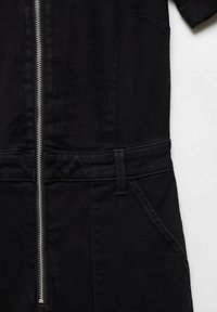 Vêtement noir avec une fermeture éclair verticale argentée, des passants de ceinture et une poche avant visible sur le côté droit sur fond blanc.