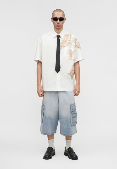 Άνδρας φορώ λευκό κοντομάνικο πουκάμισο με σχέδια ανεμιστήρων, μαύρη γραβάτα, φαρδιά ανοιχτόχρωμα τζιν σορτς cargo, μαύρα παπούτσια, λευκές κάλτσες και μαύρα γυαλιά ηλίου.