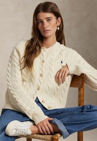 Jeune femme assise en tailleur sur une chaise en bois, portant un cardigan en maille torsadée crème, un jean bleu, des baskets blanches et des boucles d’oreilles en coquillage.
