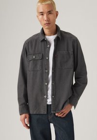 Chemise en denim gris avec deux poches poitrine, fermeture à boutons et coupe décontractée, portée par-dessus un T-shirt blanc, associée à un jean foncé.