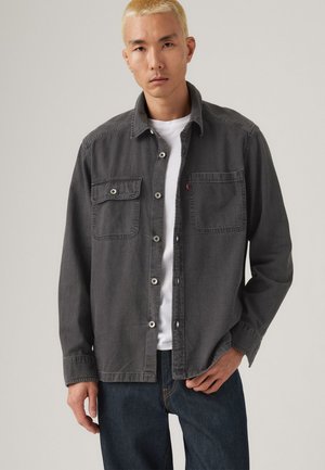 Gray denim shirt with two chest pockets, button closure, and a relaxed fit, worn over a white T-shirt, paired with dark jeans.
