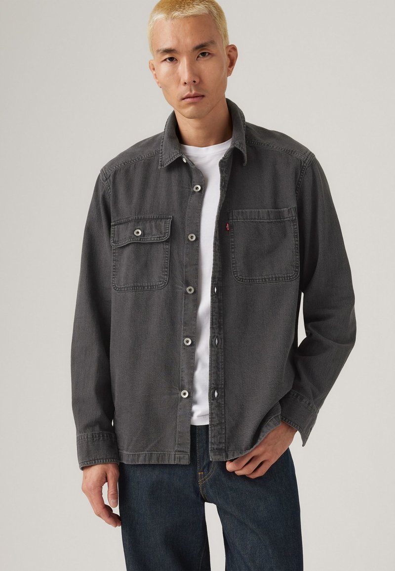 Chemise en denim gris avec deux poches poitrine, fermeture à boutons et coupe décontractée, portée par-dessus un T-shirt blanc, associée à un jean foncé.