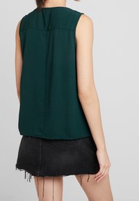 Dunkelgrünes ärmelloses Oberteil mit subtiler Textur und einem Rückenteile-Design, kombiniert mit einem schwarzen Denim-Rock mit fransigen Kanten.